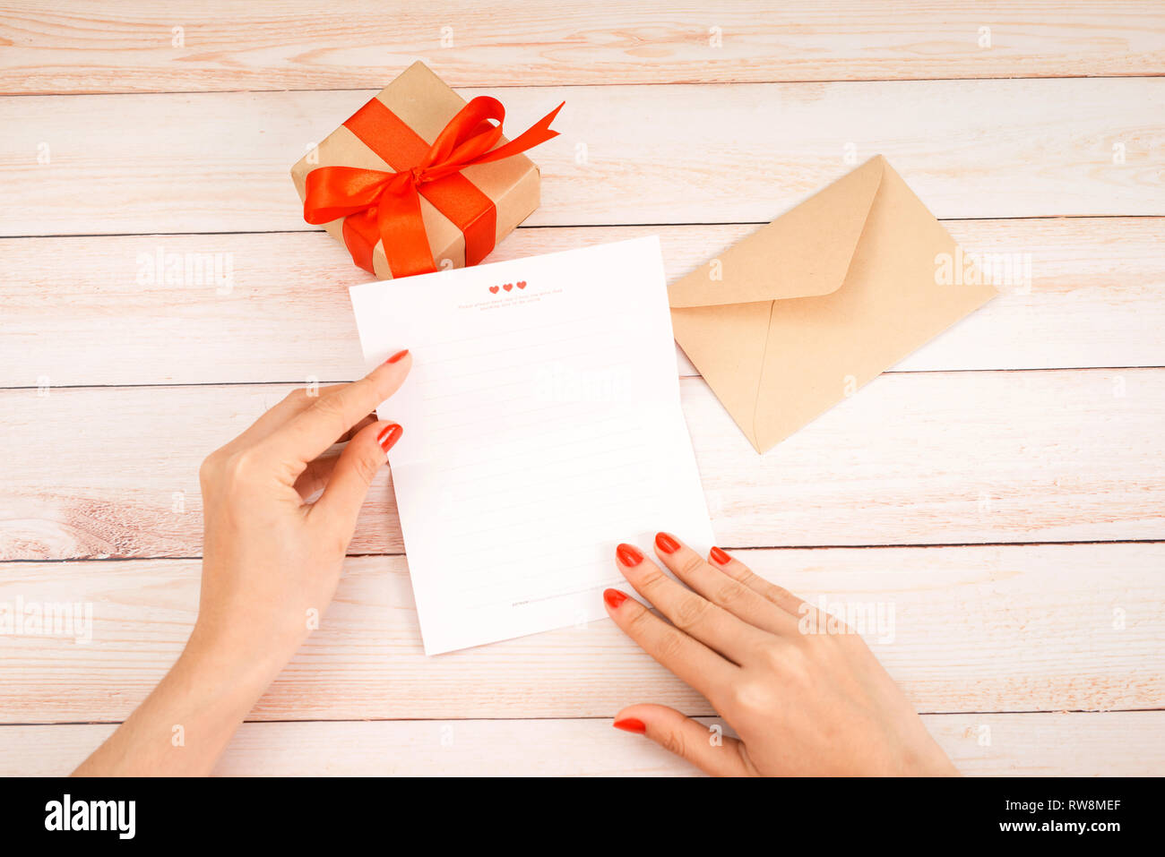 Valentine's day love letter on wooden background. Brown envelop, pink note and gift box on table.  Female hands with red nail polish Stock Photo