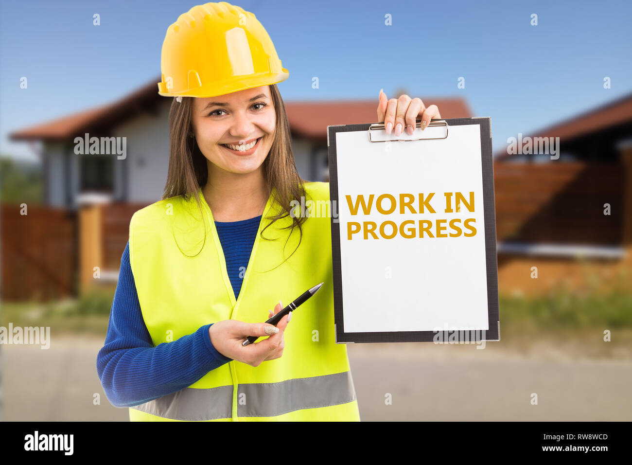 Woman builder with friendly expression holding work in progress orange text on clipboard pointing by using pen Stock Photo