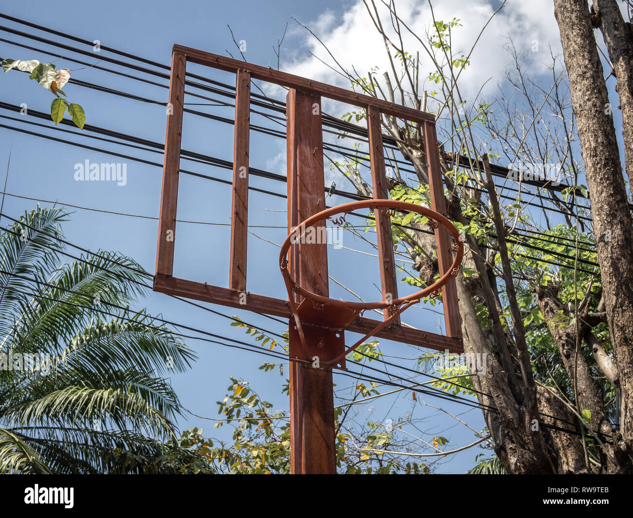 Old red basketball hoop near trees and busy electric wires on blue sky background Stock Photo