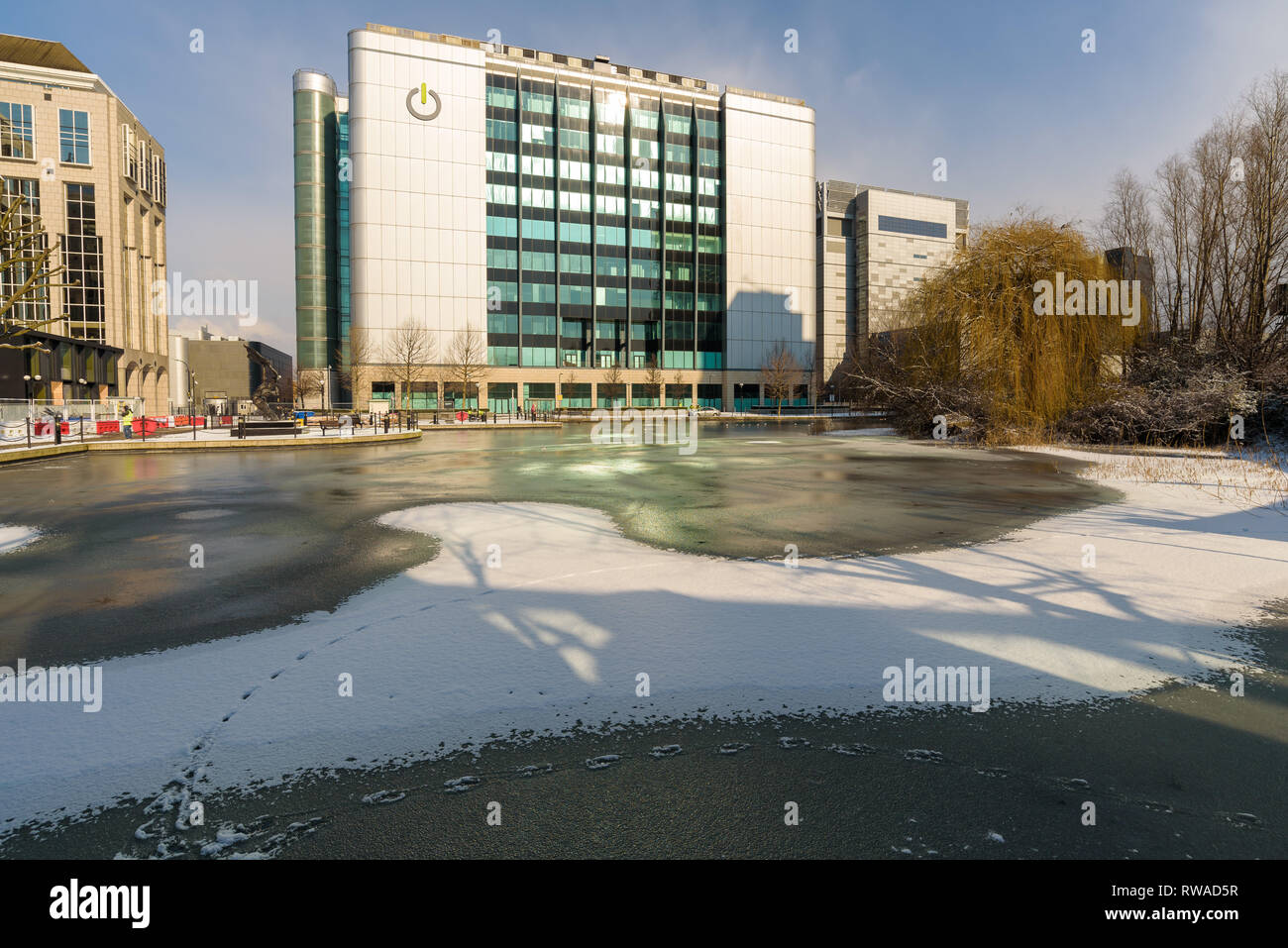 London, England - Feb 2018. Fresh snow covers the frozen pond next to East India DLR station in Poplar. In the background the Global Switch building. Stock Photo