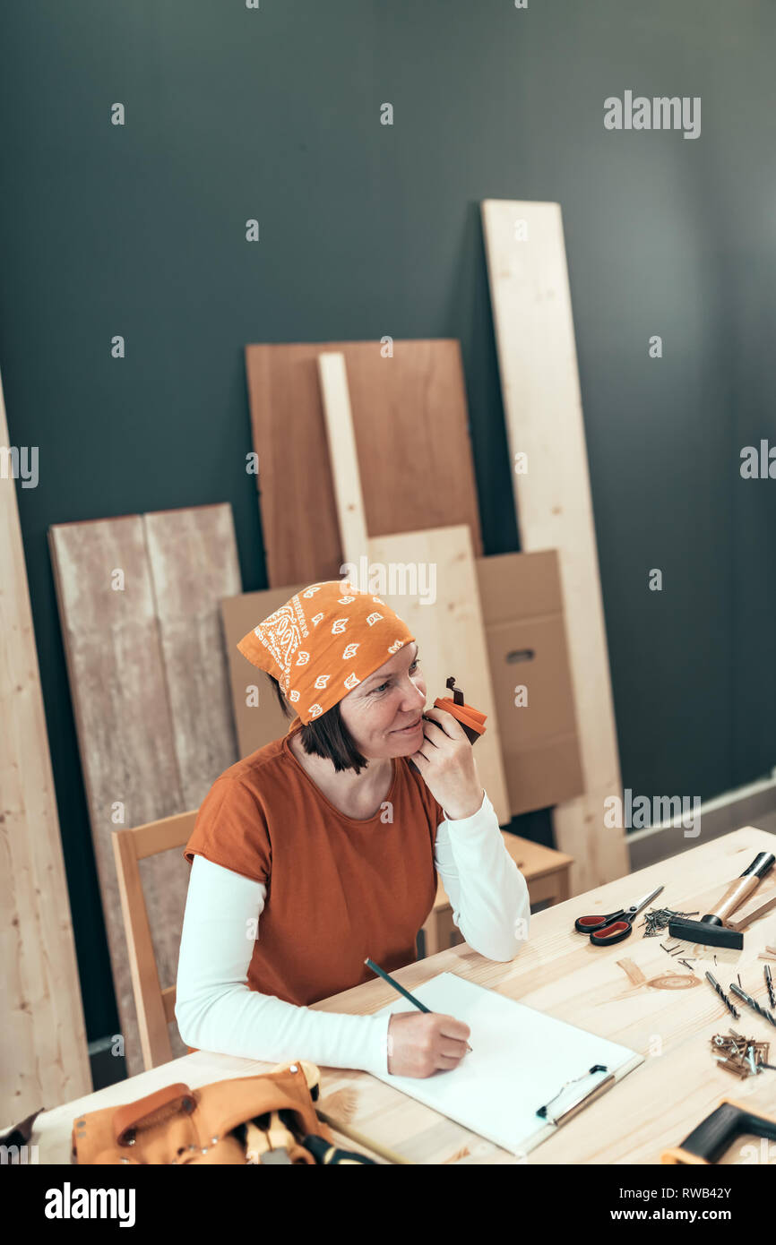 Female carpenter is drinking coffee and planning DIY project in her small business woodwork workshop Stock Photo