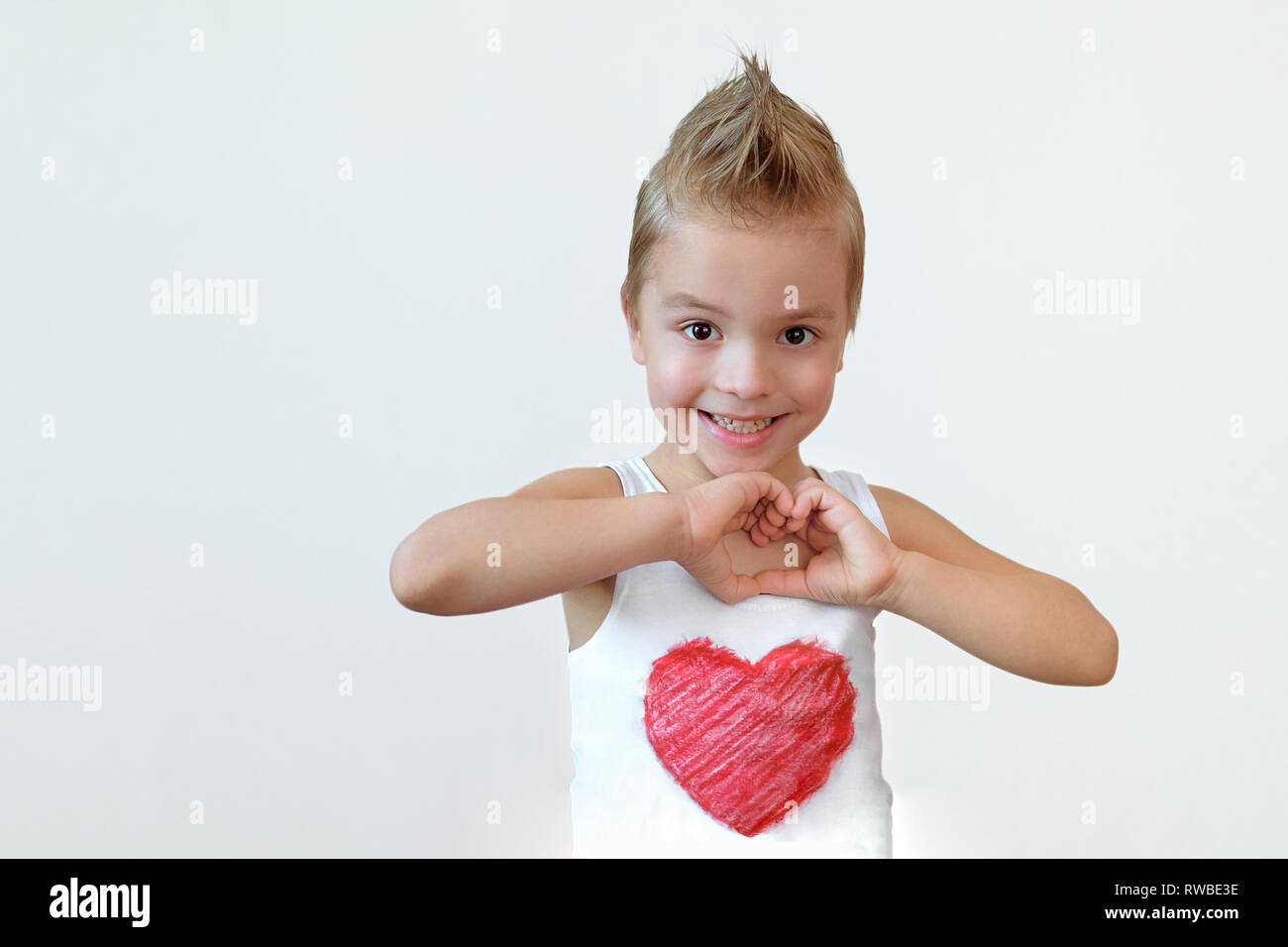 Lovely child boy smiling shows heart sign. Portrait kid blond 6 year with smile making hands gesture love. Studio isolated white background. Stock Photo