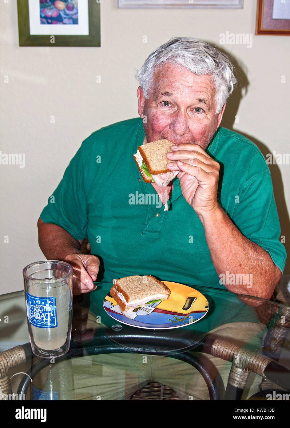 senior man eating sandwich; biting, ham; cheese; lettuce; whole wheat bread; food; nutritious; glass table; lemonade; insulated glass; meal; home, ver Stock Photo