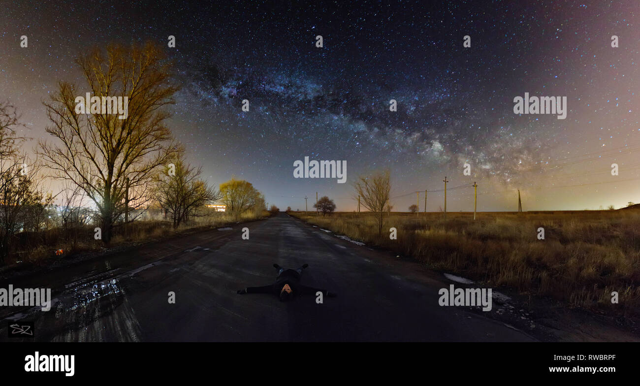 The guy lying below the night starry sky. Man lying on asphalted road and see into the night sky. Watching milky way. Stock Photo