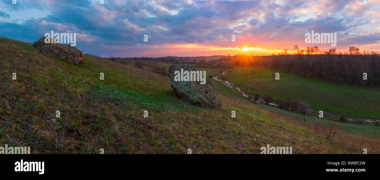 A skewed valley panoramic view of nature spring landscape. Spring landscape with big stone under the amber cloudy sunset. Valley with river Stock Photo