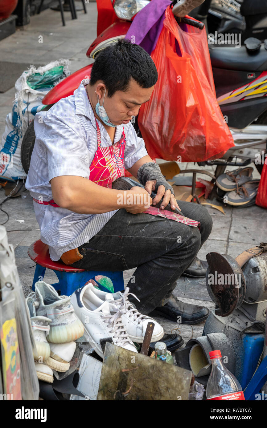 Man working as a cobbler , repairing shoes, on the pavement in the Old Quarter of Hanoi, Vietnam, Asia Stock Photo