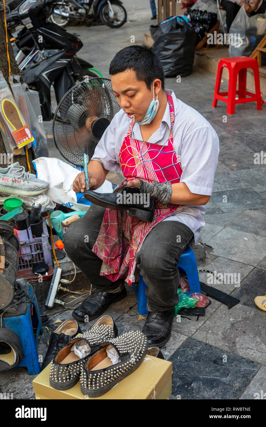Man working as a cobbler , repairing shoes, on the pavement in the Old Quarter of Hanoi, Vietnam, Asia Stock Photo