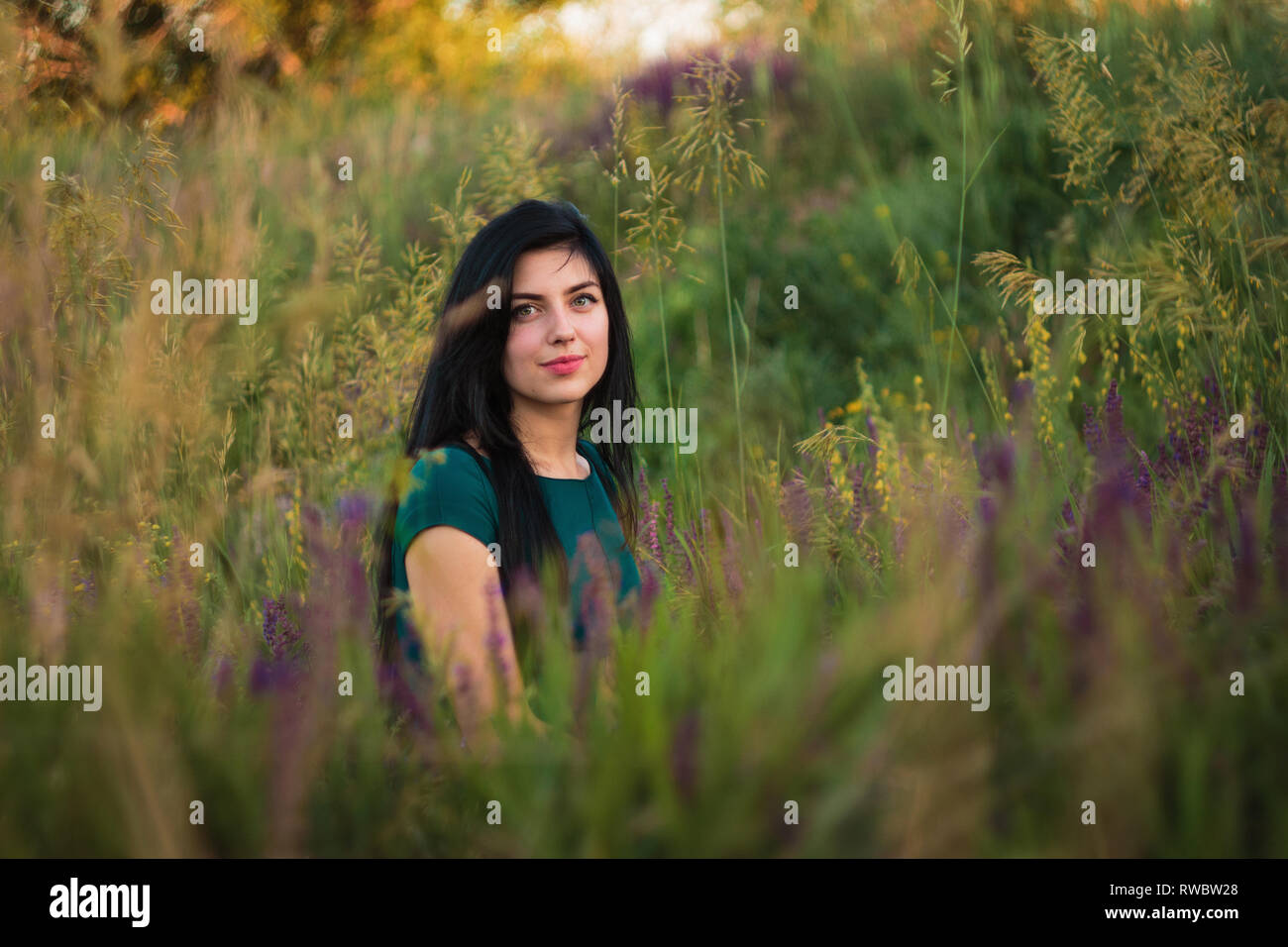 Beautiful young girl with black hair and green eyes sitting in the grass and violet flowers on green background in Dnipropetrovsk region, Ukraine Stock Photo