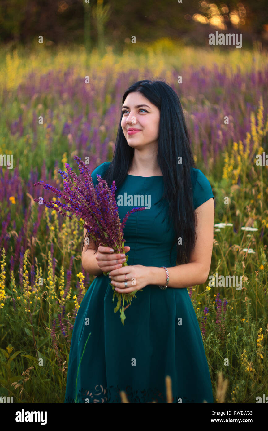 Beautiful happy young girl dressed in marine-colored dress staying at the flowers field and taking bouquet of violet flowers Stock Photo