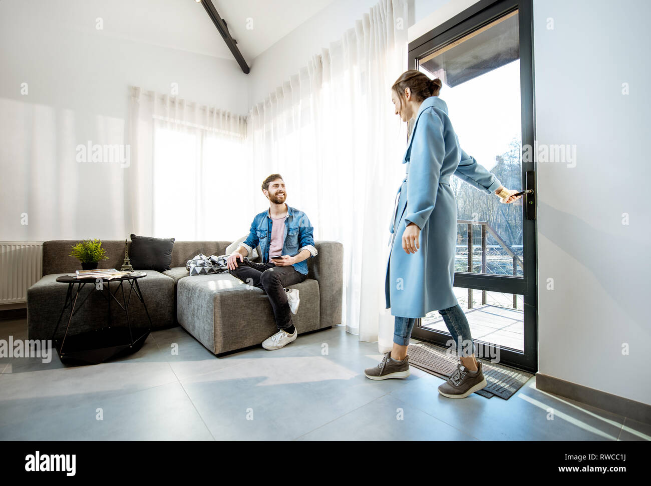 Young woman in blue coat coming home and meeting her husband on the couch in the living room Stock Photo