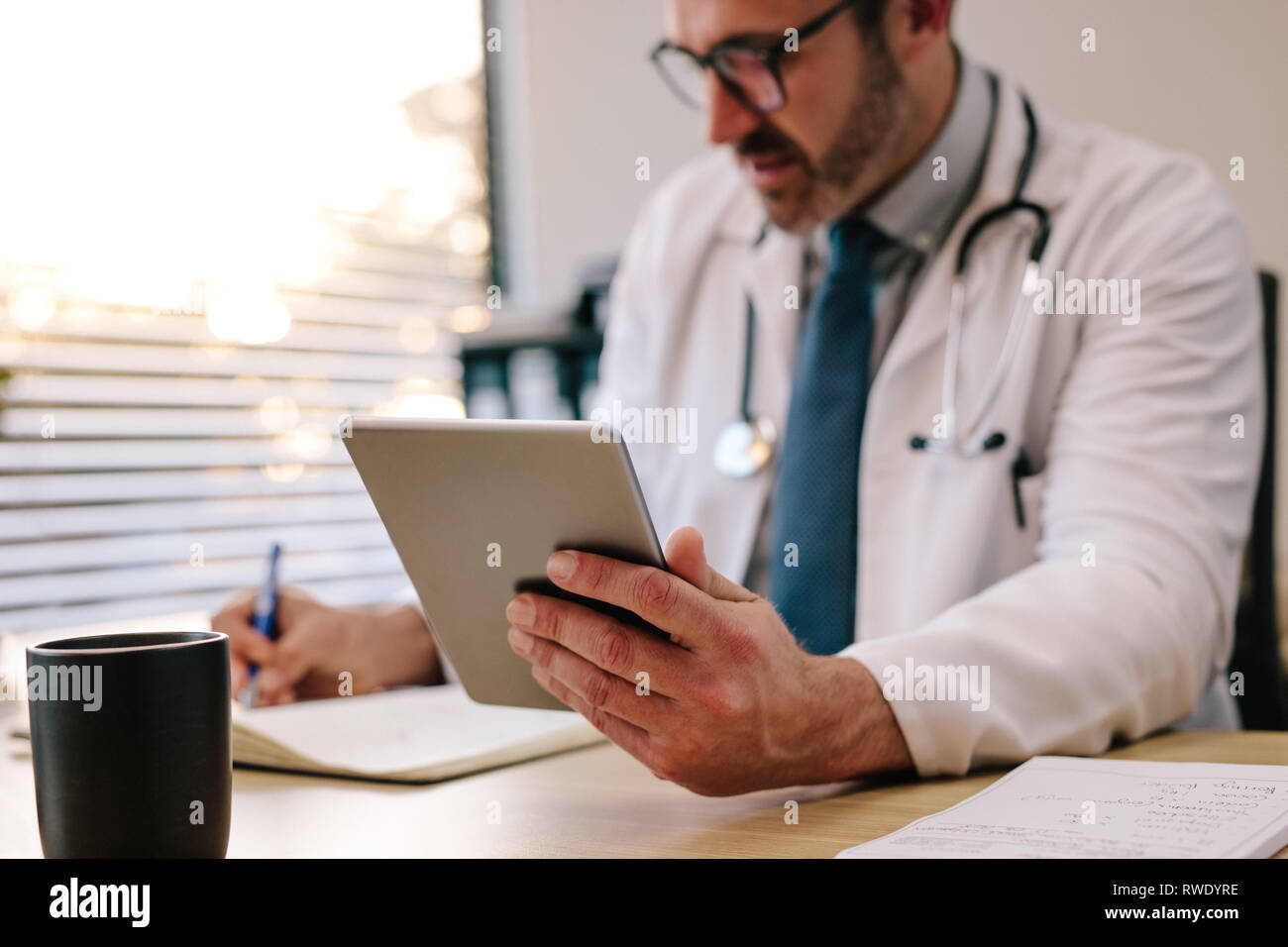 Mature male doctor holding a digital tablet making notes in his diary while sitting in his office. Doctor sitting at his desk with tablet computer and Stock Photo