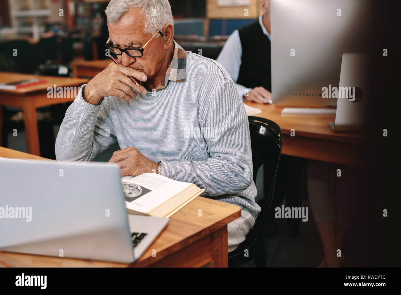 Elderly gentleman reading a book sitting in classroom with a laptop in front. Senior man learning in a university classroom. Stock Photo