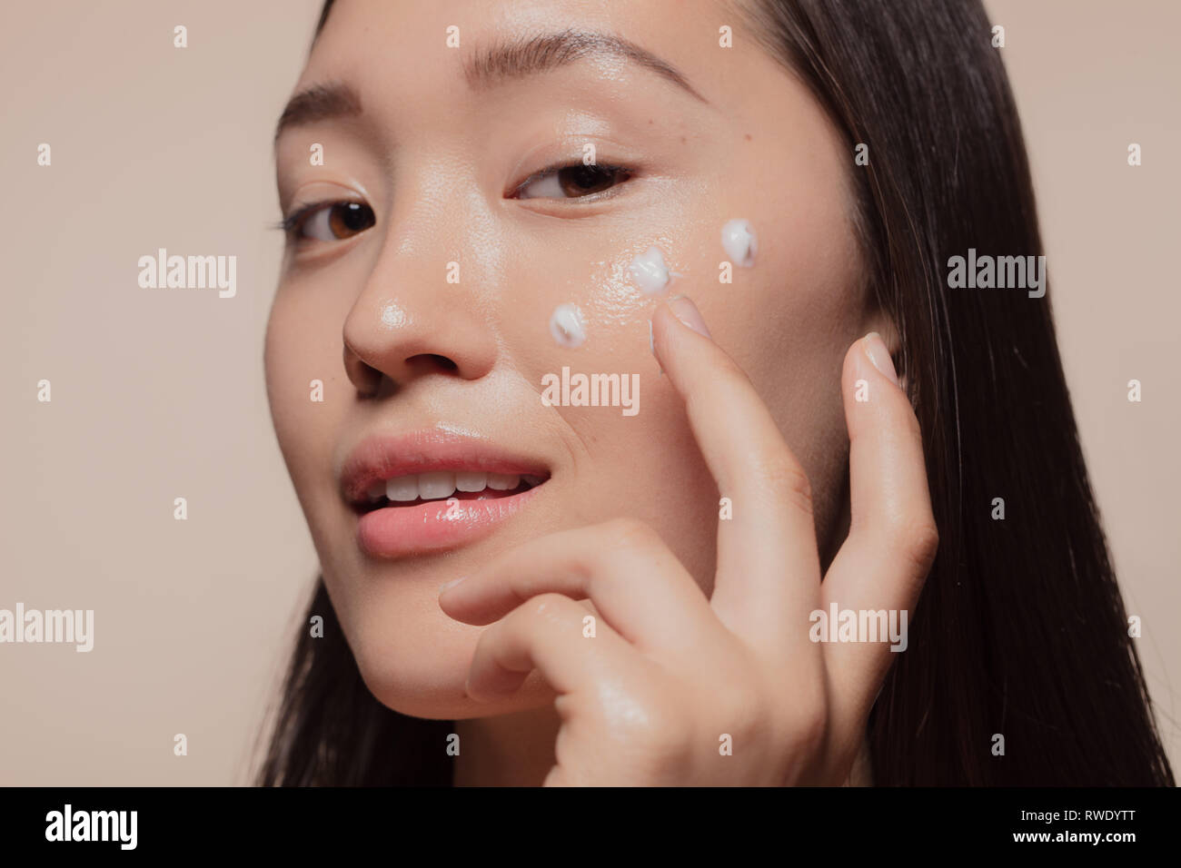 Close up of a young woman applying moisturizer to her face. Asian woman looking happy while following skincare regime. Stock Photo
