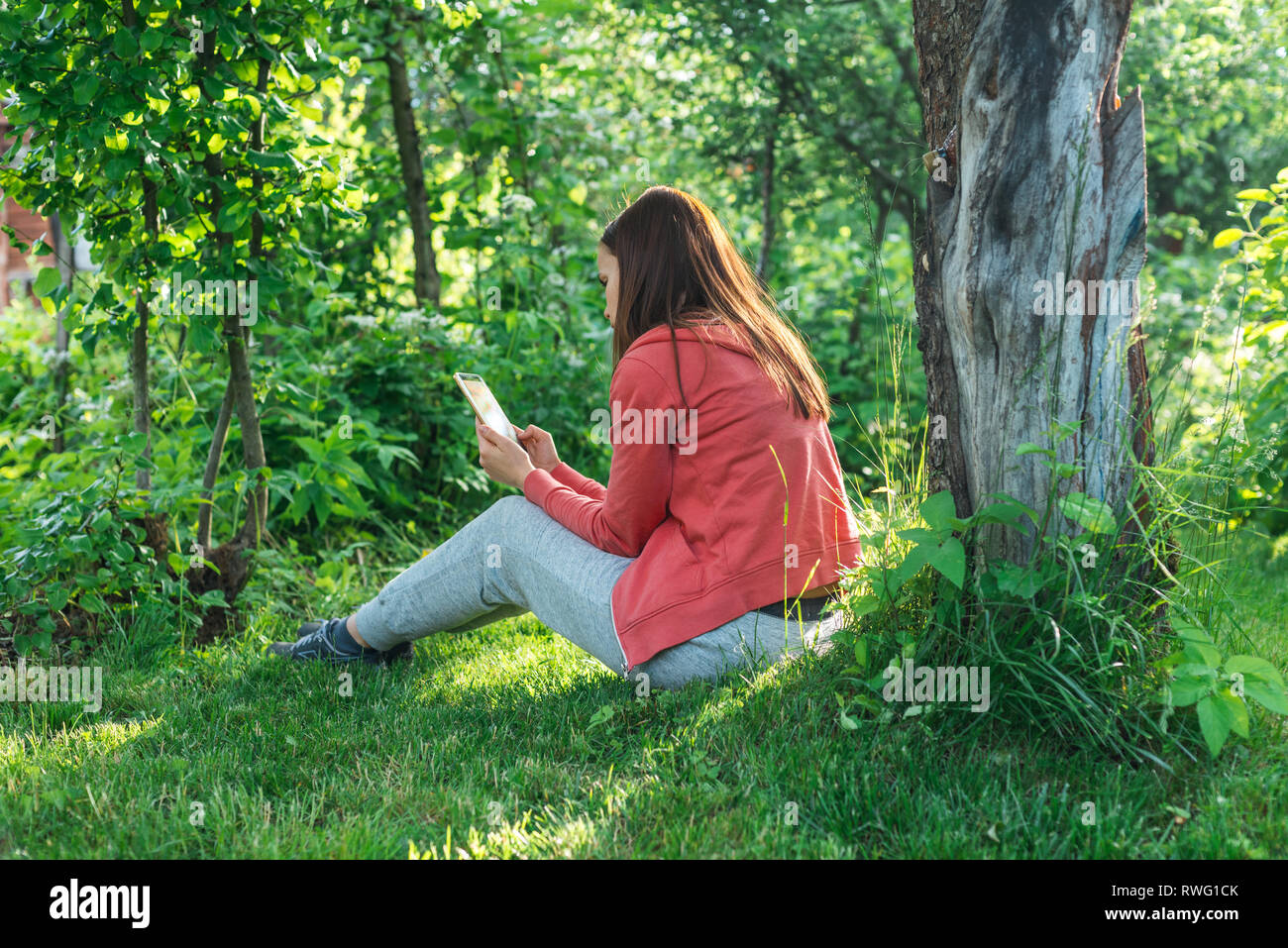 Girl in a sporty pink jacket and gray pants sits on a green spring lawn near a tree and reads on the phone Stock Photo