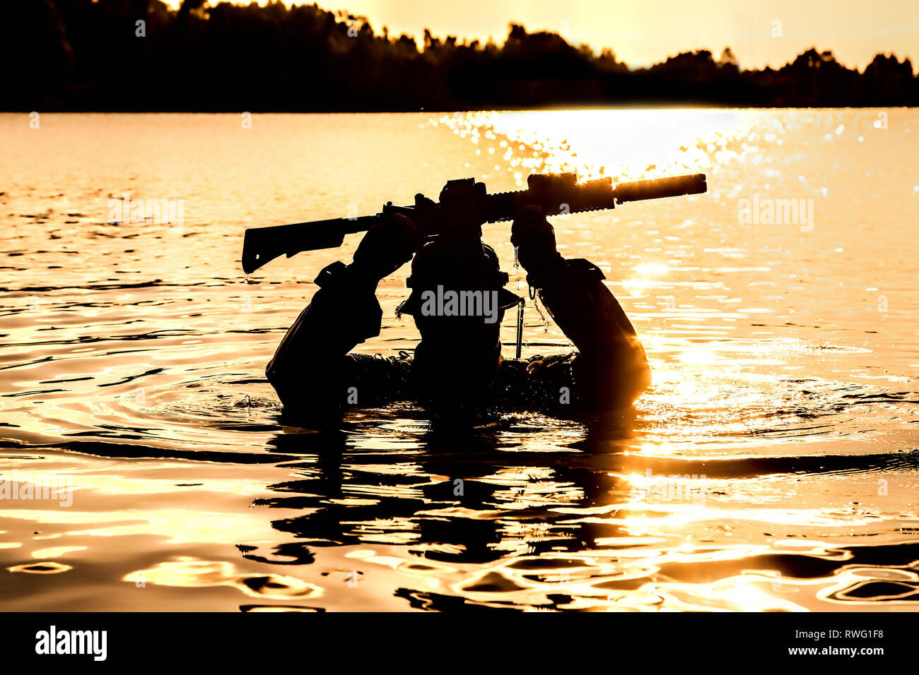 Silhouette of a special forces soldier holding weapon above head while crossing a river. Stock Photo