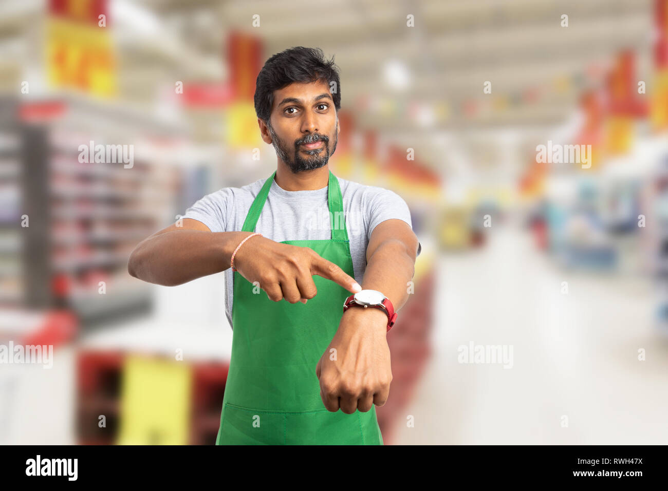 Upset indian male supermarket employee or manager with serious expression pointing with index finger at watch on wrist as you are late for work concep Stock Photo