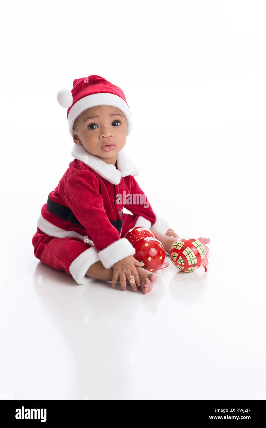 Six Month old baby girl wearing a Santa Claus costume. She is sitting on a white, seamless background with Christmas ornaments. Stock Photo