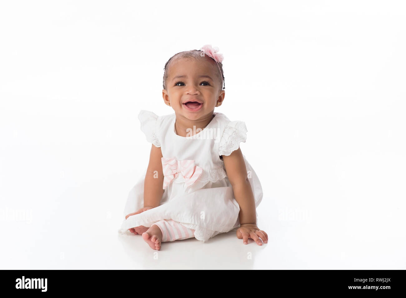 A smiling six month old baby girl wearing a white dress. She is sitting on a white, seamless background. Stock Photo