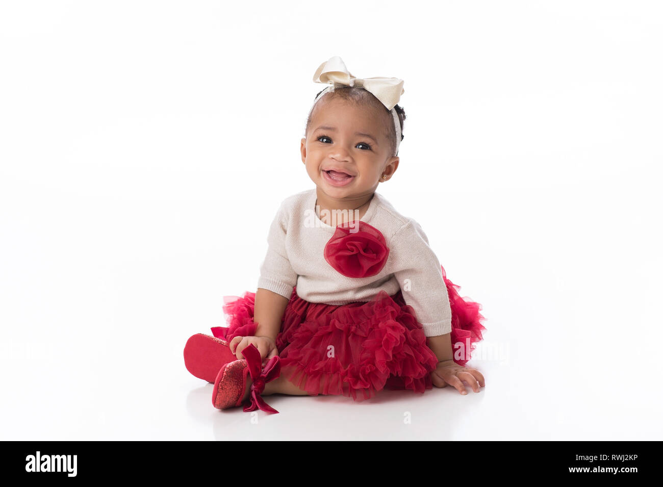A smiling six month old baby girl wearing a red tutu. She is sitting on a white, seamless background. Stock Photo