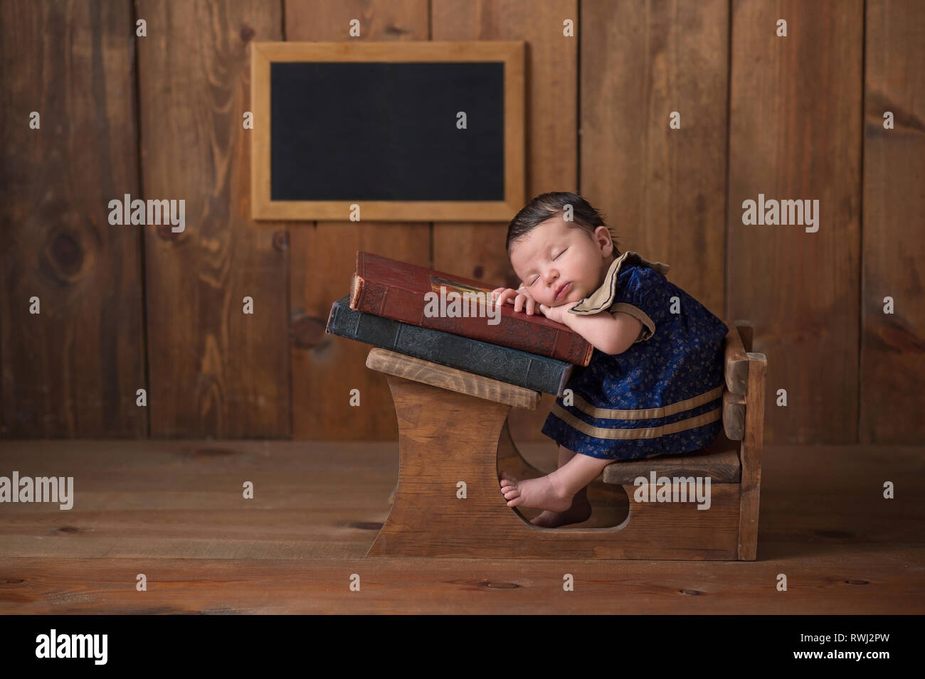 One week old newborn baby girl wearing a dress and sleeping on a stack of vintage books at a tiny school desk. Stock Photo