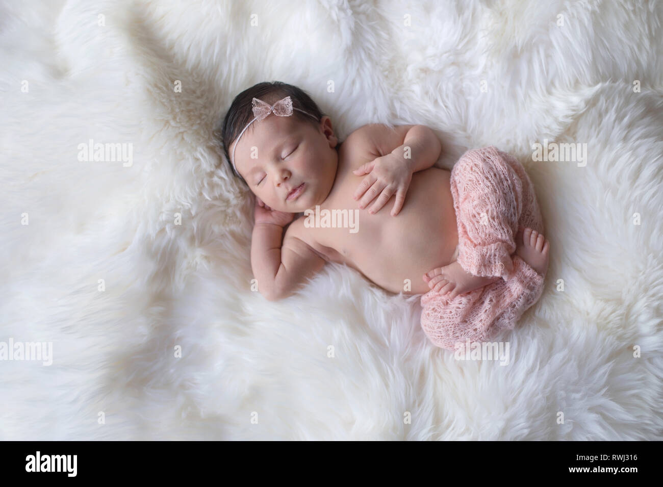 Sleeping, week old newborn baby girl wearing light pink, knitted pants. Shot in the studio on a white sheepskin rug. Stock Photo