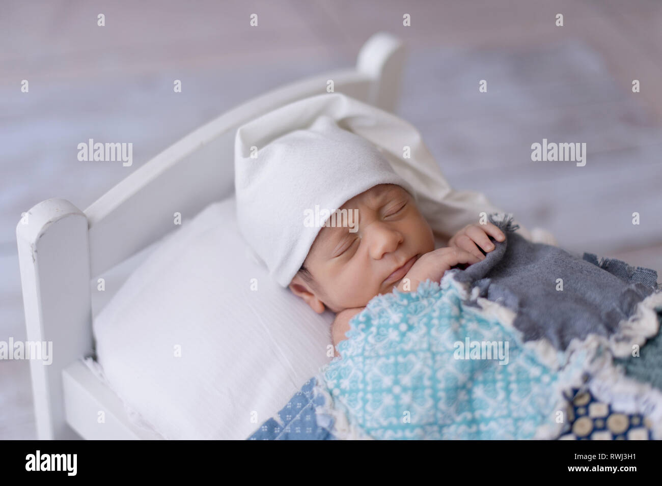 One week old newborn baby boy wearing a white sleeping cap. He is sleeping on a tiny, white bed and covered with a blue, patchwork quilt. Stock Photo