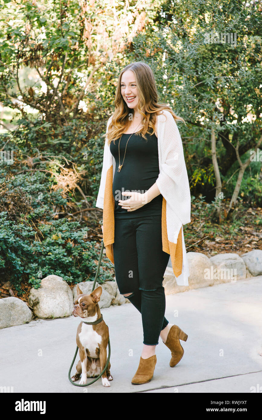 An expectant mother holds her pregnant belly while walking her dog outdoors in the park Stock Photo
