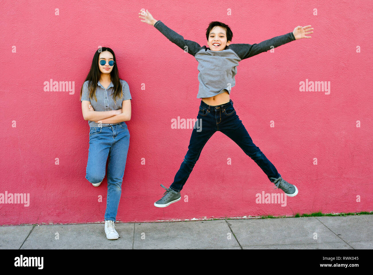 A little boy tries to impress his older sister by jumping high.  She stands against an urban wall unimpressed Stock Photo