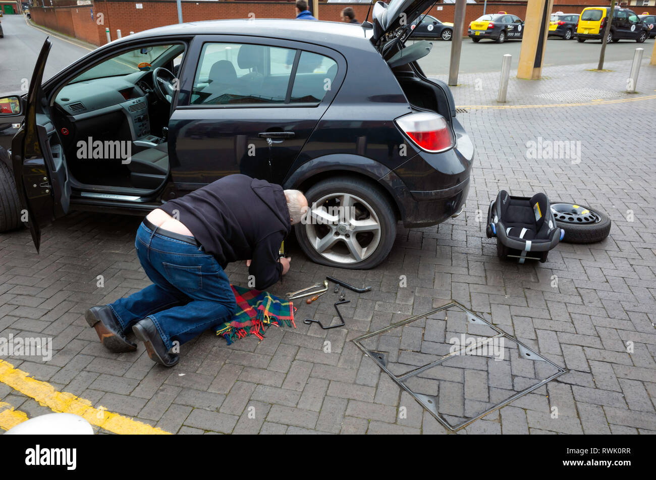 Elderly man kneeling beside his car preparing to change the rear wheel with its punctured tyre in a town centre Stock Photo