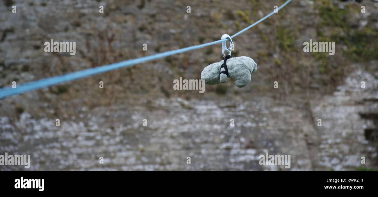 A small green teddy bear on a zipwire with a stone wall in the