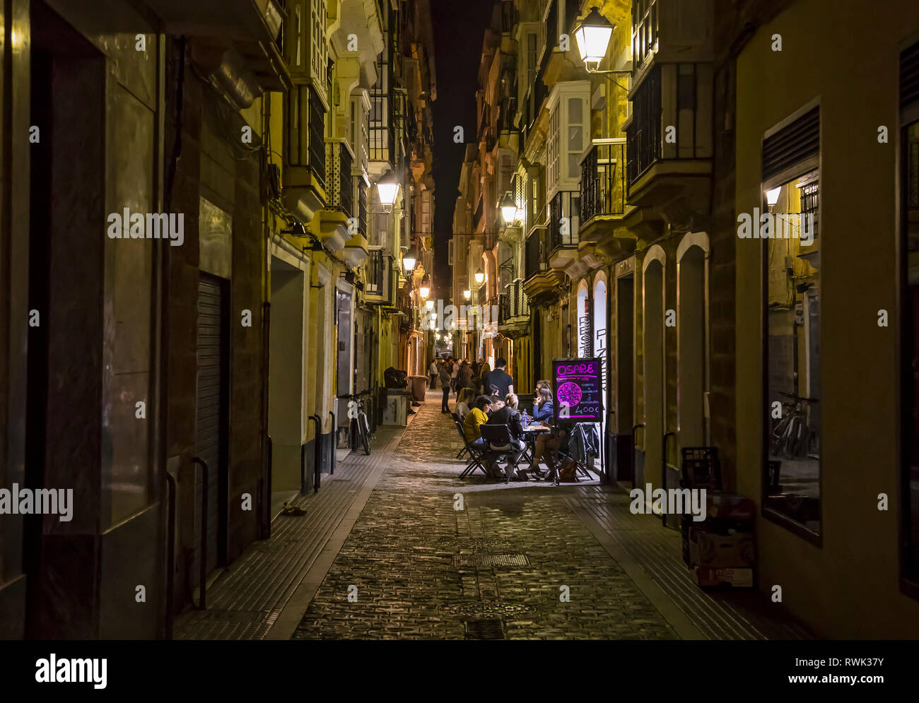 Patrons sit at tables on a patio flooded with light in a quiet, narrow street at night; Cadiz, Andalusia, Spain Stock Photo