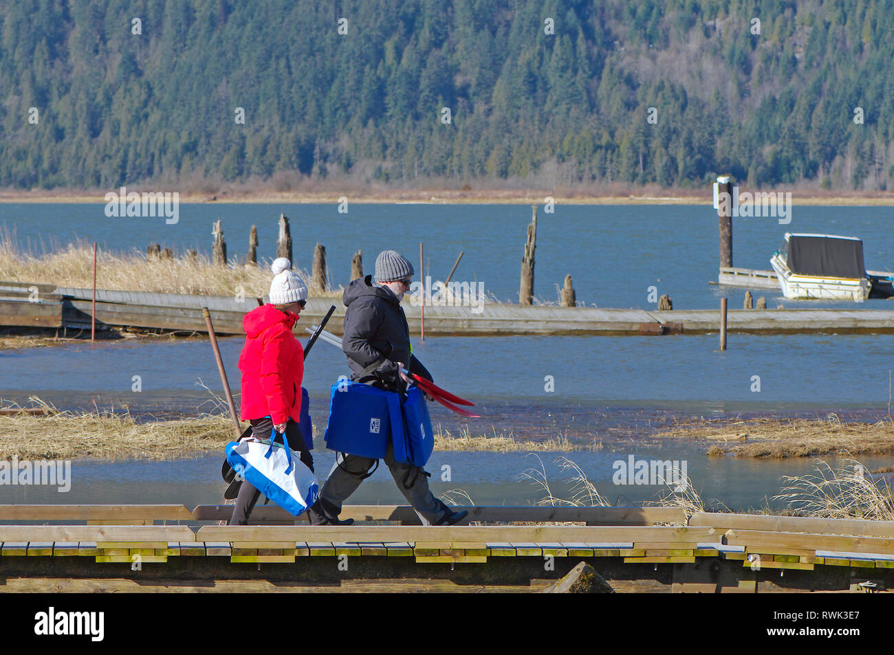 Middle aged couple in winter clothing walking with life jackets and kayak paddles along a pier in Grant Narrows Regional Park, B. C., Canada. Stock Photo
