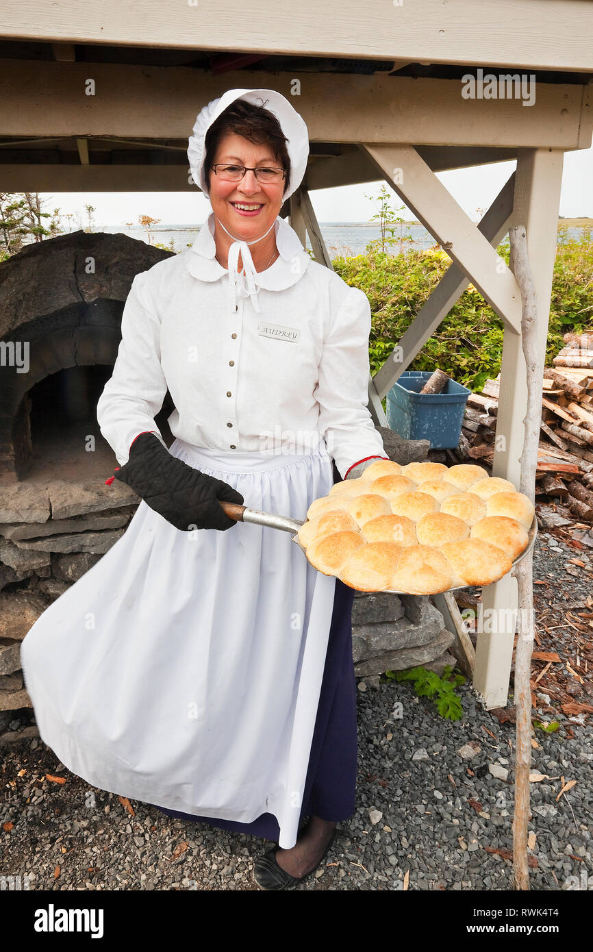 Parks Canada interpreter in period costume holding a pan of freshly baked bread that she just removed from a traditional, outdoor brick and stone oven behind her. Port au Choix National Historic Site, Port au Choix, Newfoundland, Canada Stock Photo