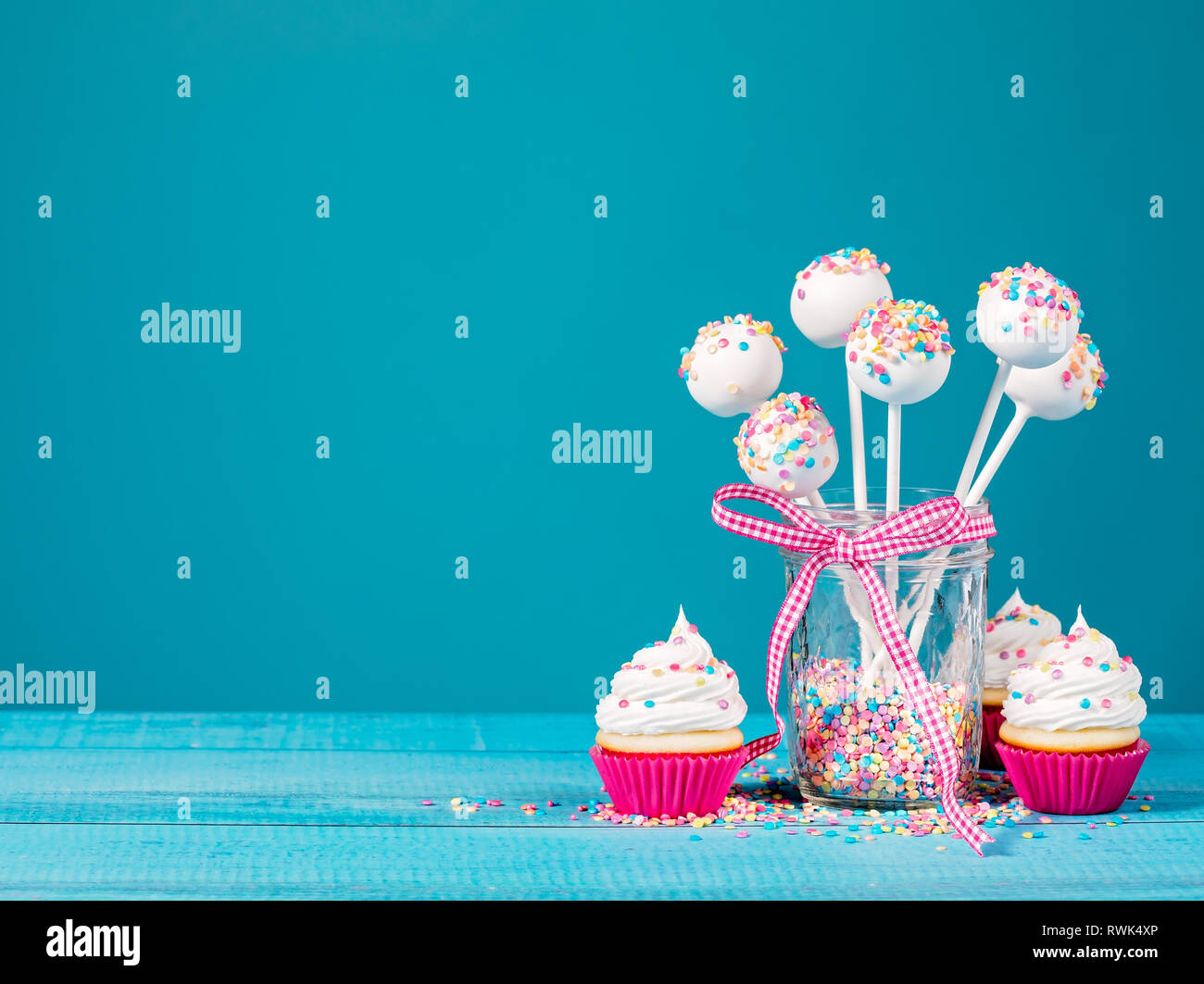 Vanilla cake pops with colorful sprinkles and cupcakes on a blue background. Stock Photo