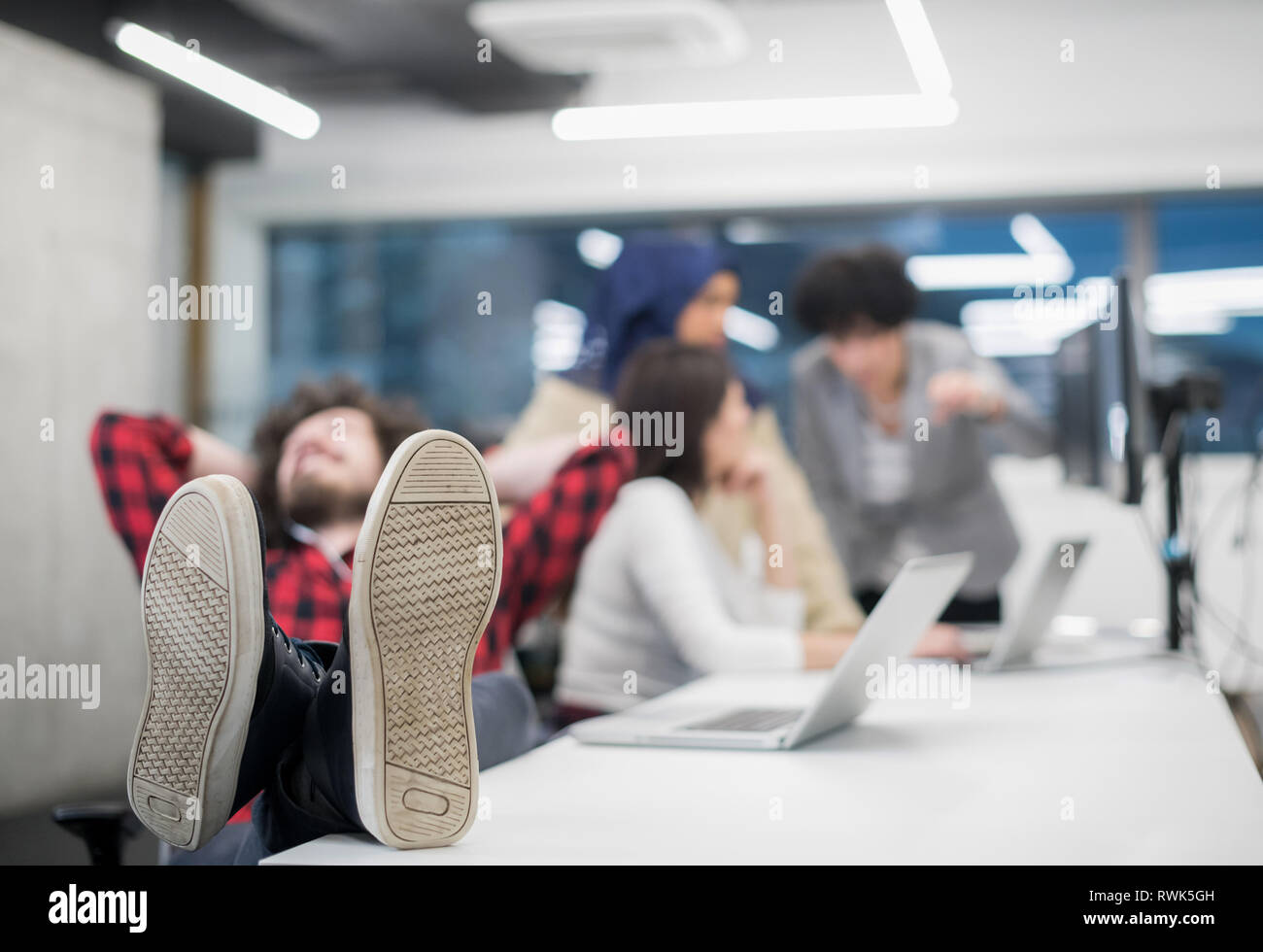young software developer resting with legs on desk while his multiethnics business team writing programming code in the background at startup office Stock Photo