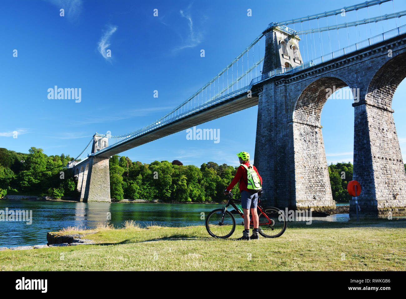 A cyclist admires the mighty Menai Suspension Bridge which joins Anglesey with the Welsh mainland over the Menai Strait. Stock Photo