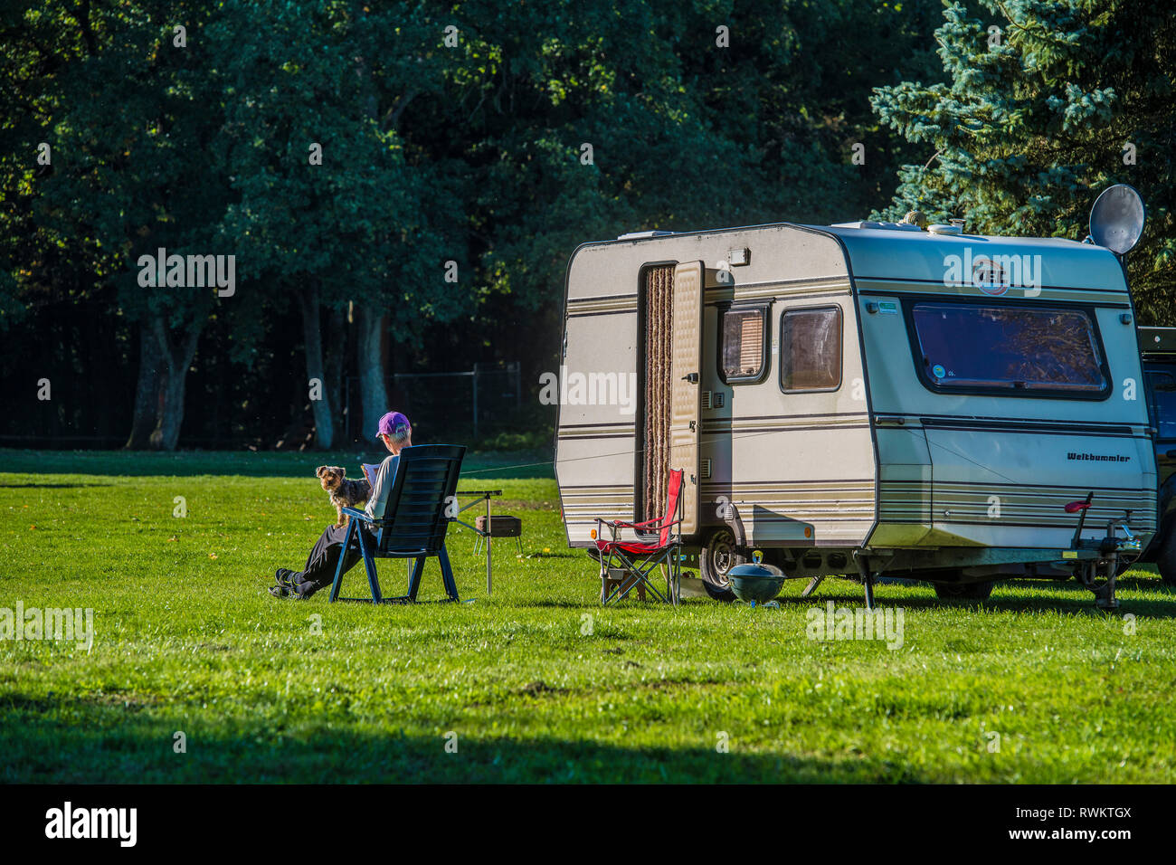 GERMANY, RHINELAND PALATINATE; FERSCHWEILER, Man reading with dog on his lap enjoying the autumn sun on a clearing in front of their caravan Stock Photo