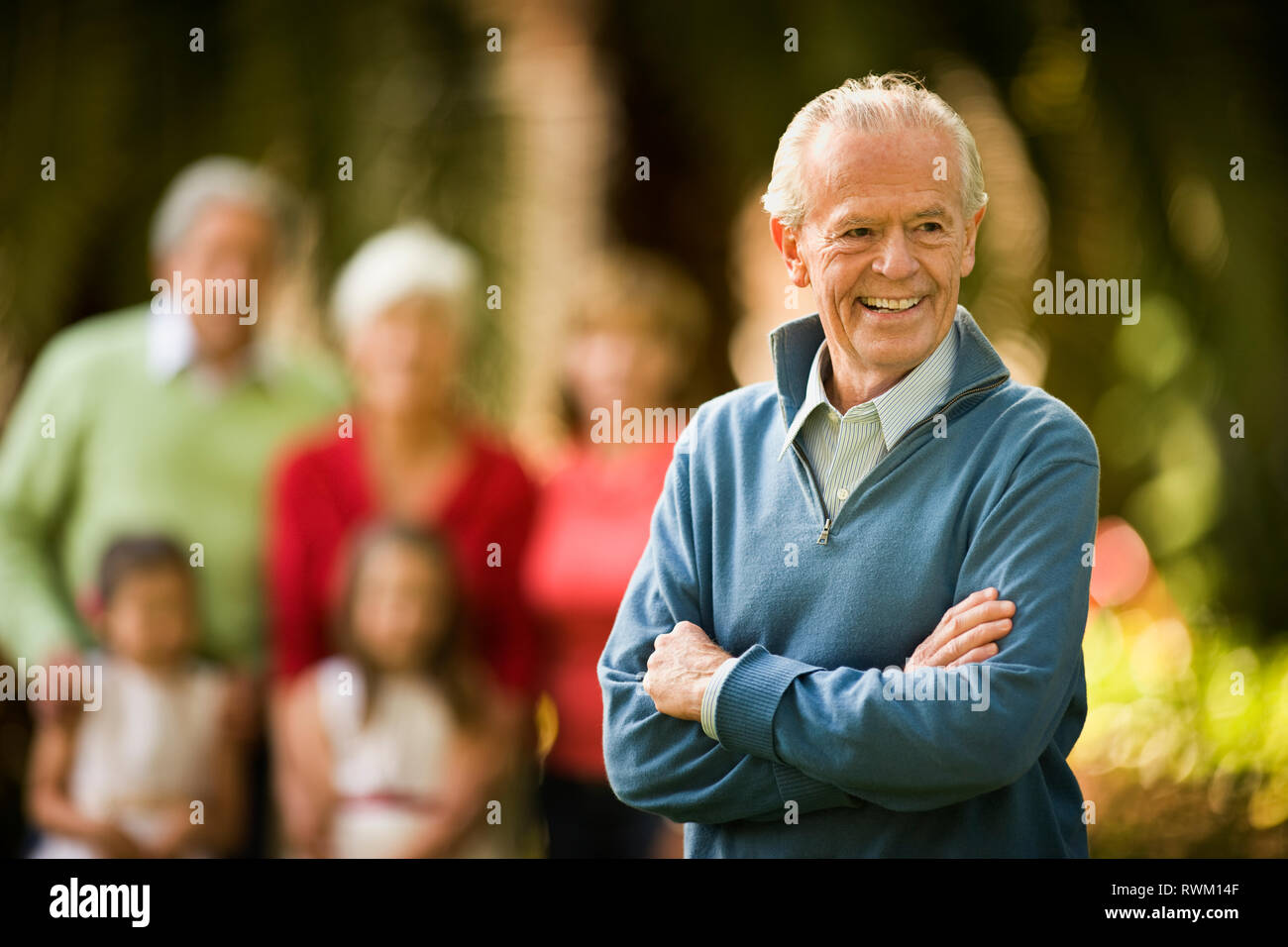 Senior man smiles proudly and stands with his arms crossed as he poses for a portrait with his family in the background. Stock Photo