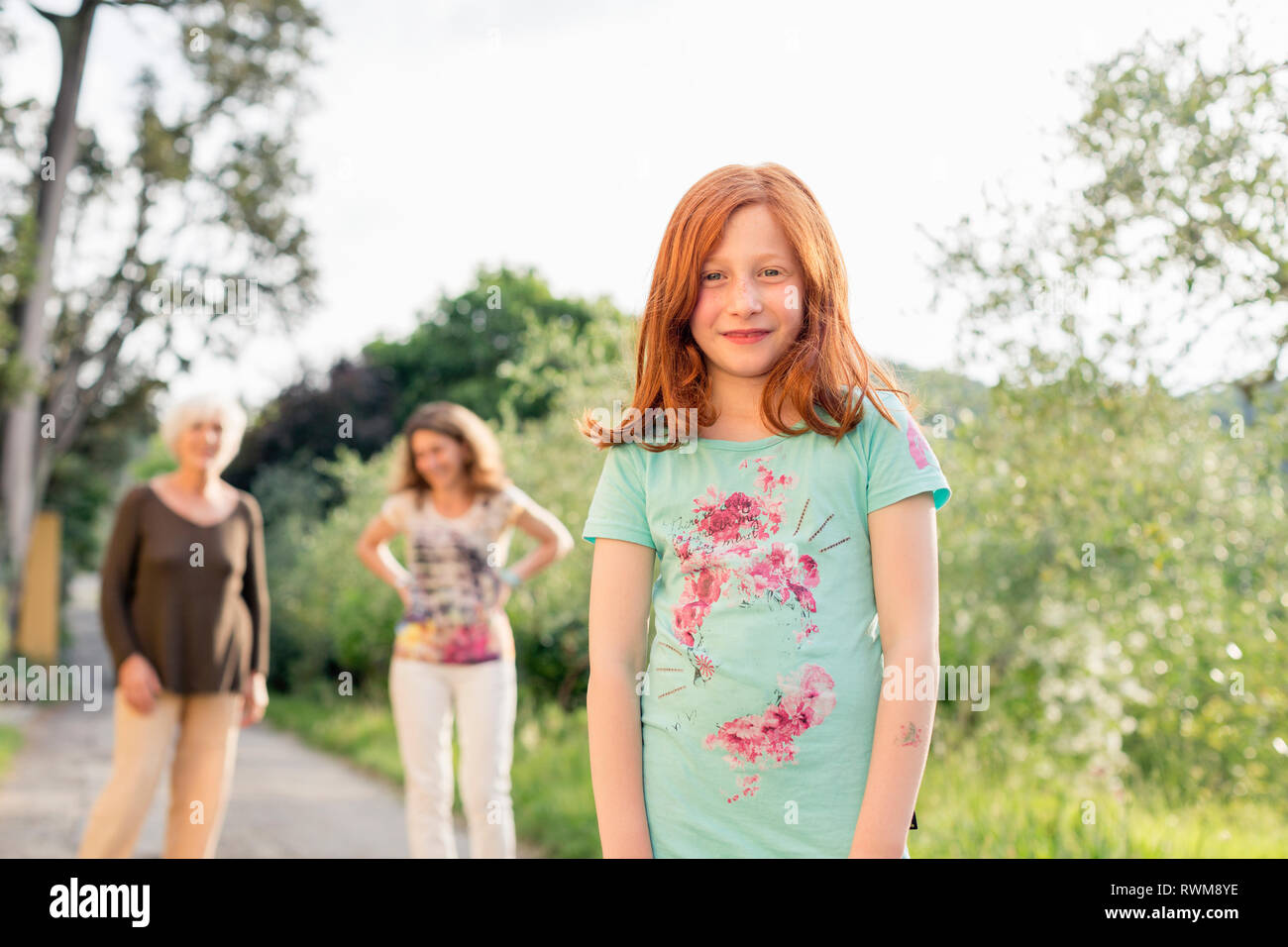 Red haired girl on rural road with mother and grandmother in background, portrait Stock Photo