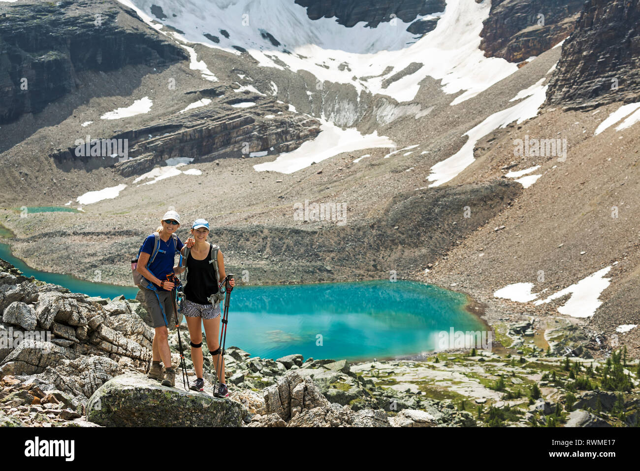 Two female hikers standing in a large rocky area with a colourful alpine lake and mountain cliffs with snow in the background Stock Photo