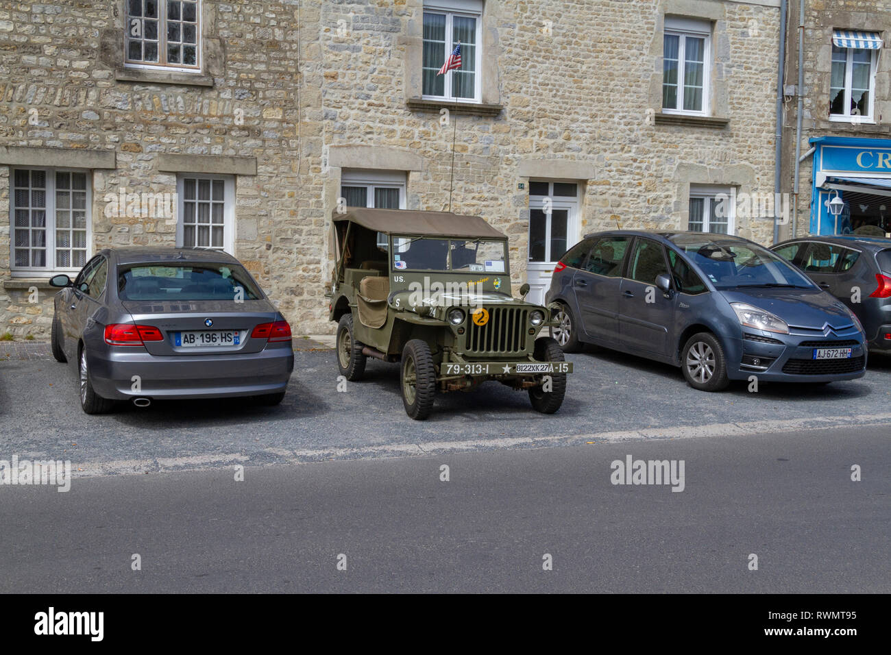 A US Army World War Two jeep parked between two modern cars in Sainte-Marie-du-Mont, Normandy, France. Stock Photo