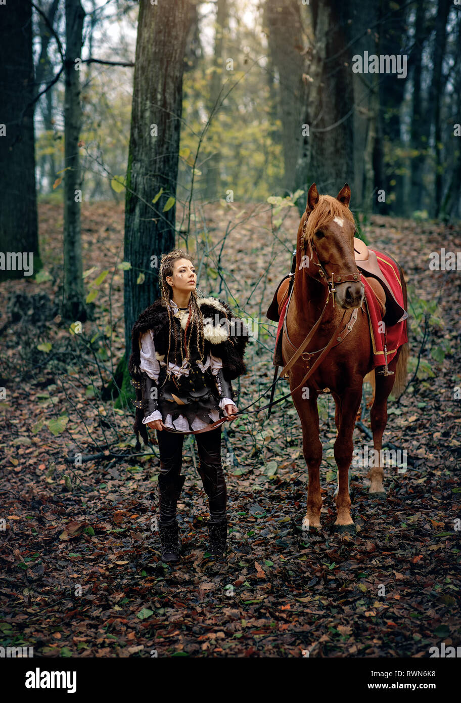 Warrior Woman and a horse in the woods. Scandinavian viking with her horse in traditional clothes with fur collar, war makeup, forest. Reconstruction  Stock Photo
