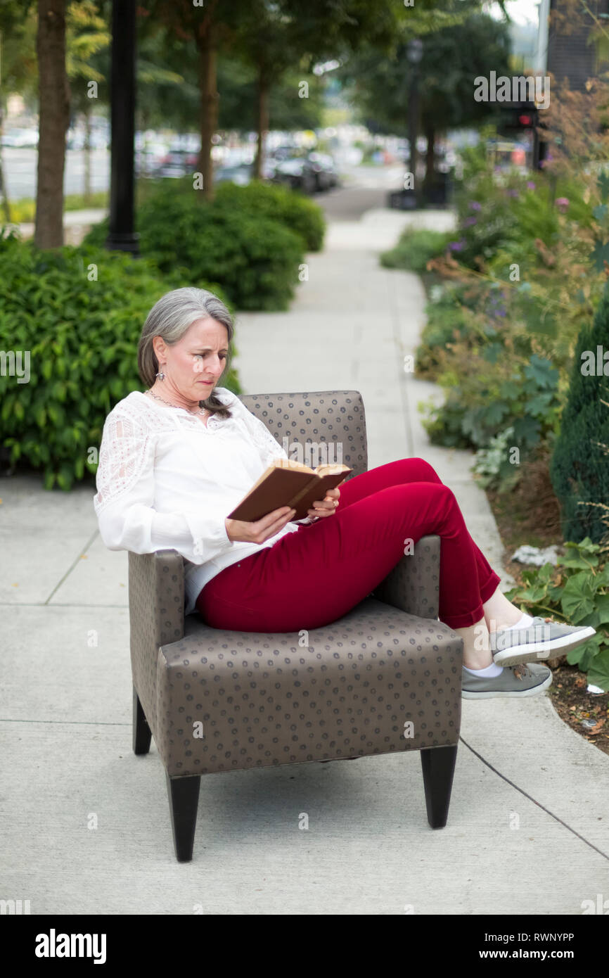 A mature woman sits reading a book in an armchair on an outdoor walkway; Bothell, Washington, United States of America Stock Photo
