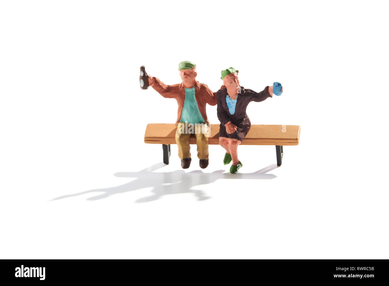 Miniature couple sitting on a bench celebrating toasting the camera saying cheers as they lift their beers in the air with shadow on white Stock Photo