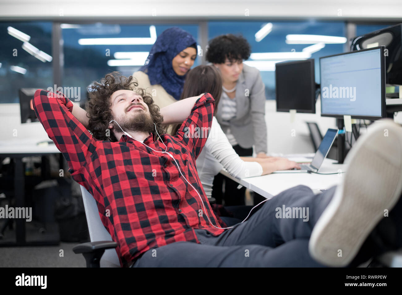 young software developer resting with legs on desk while his multiethnics business team writing programming code in the background at startup office Stock Photo