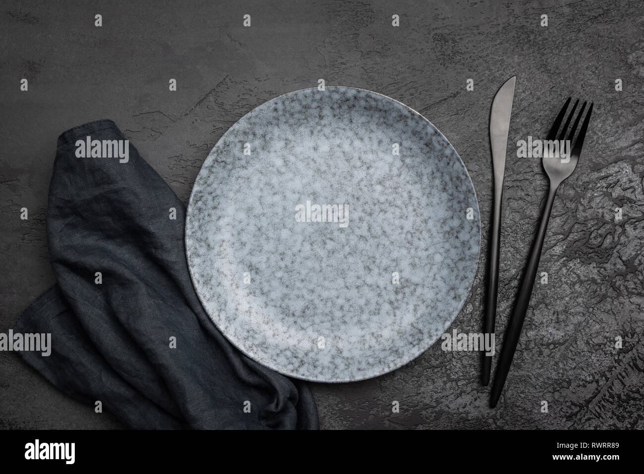 Stylish table setting with black cutlery. Empty plate, table textile and black cutlery on black concrete background. Top view. Restaurant, menu, cafe  Stock Photo