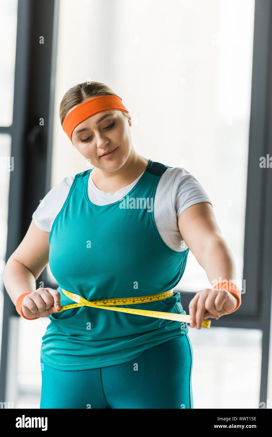 plus size girl looking at measuring tape while measuring waist in gym ...