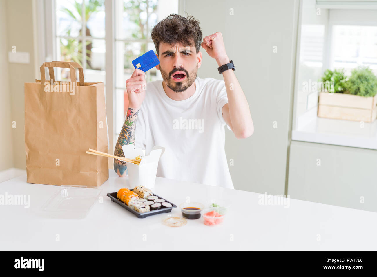 Young man eating asian sushi from home delivery using credit card as payment annoyed and frustrated shouting with anger, crazy and yelling with raised Stock Photo