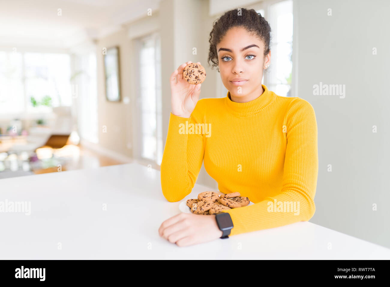 Young african american girl eating chocolate chips cookies as sweet snack with a confident expression on smart face thinking serious Stock Photo