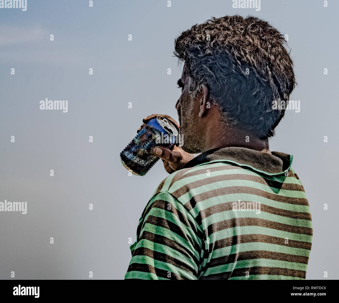 Middle Aged Fisherman of South Asian Origin, wearing striped tshirt is exhausted and quenching his thirst by drinking beer/cold drink on hot sunny day Stock Photo